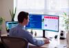 man in a gray dress shirt sitting on a chair in front of a computer monitor