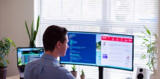 man in a gray dress shirt sitting on a chair in front of a computer monitor