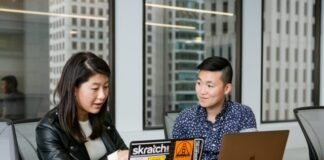 woman in black leather jacket points to laptop covered in stickers, sitting beside person in blue shirt with silver laptop