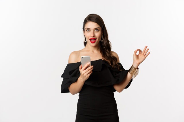 online-shopping-concept-woman-trendy-black-dress-makeup-showing-okay-sign-approval-using-mobile-phone-app-white-background