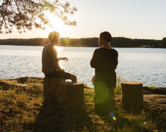 man in black jacket standing beside body of water during sunset