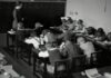 grayscale photography of teacher standing near chalkboard and children sitting on chairs