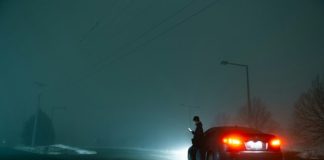 man in black jacket and black pants standing beside red car during night time